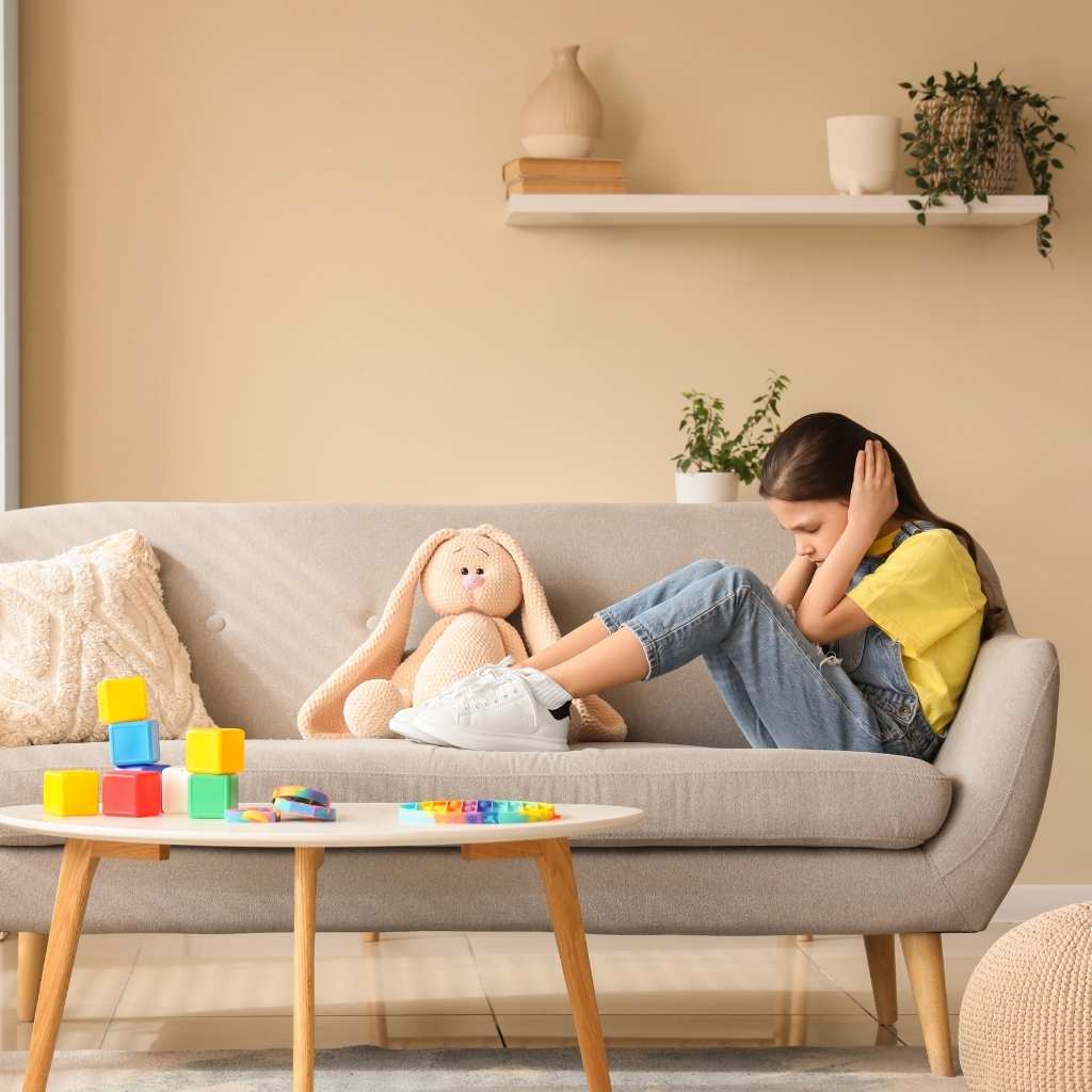 A child on a sofa covering their ears. The image reflects how autism level 1 is often misunderstood because signs can seem subtle.