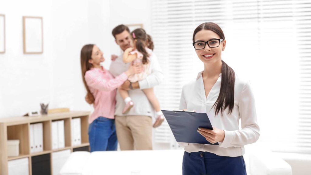 Clinician with clipboard smiles in foreground; parents hold young child in background during autism assessment visit