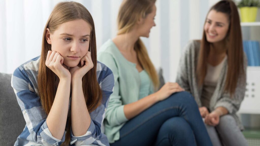 Quiet teen on a couch looking down while classmates talk nearby, showing the challenge of joining in and the need for support and understanding in autistic adolescence.